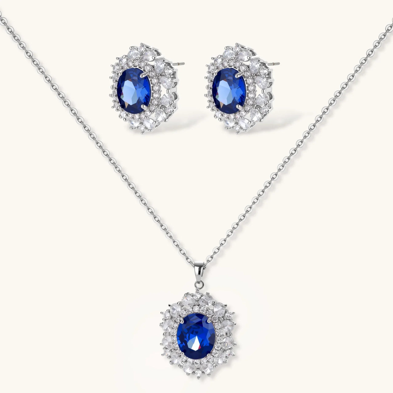 White gold plated jewelry set with blue cubic zirconia centers surrounded by clear crystal halos on cream background.
