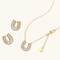 Gold-plated horseshoe necklace and earring set with baguette and round crystals arranged in geometric pattern on white background.